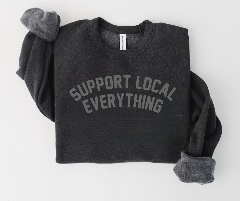 May include: Dark gray sweatshirt with the text "SUPPORT LOCAL EVERYTHING" in gray block letters. The sweatshirt has long sleeves with gray cuffs and a crew neck. The garment is made of a soft material.