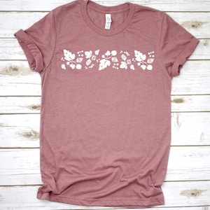 May include: A mauve colored t-shirt with a white floral design on the front. The design features leaves and berries.