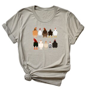 May include: A light grey t-shirt featuring a graphic of a line of chickens in various colours and breeds. The chickens are all facing forward with their wings tucked in.