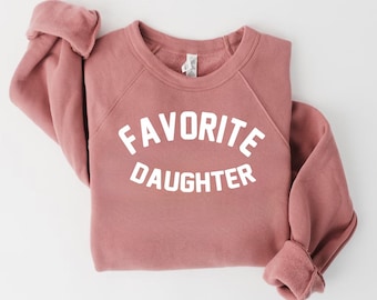Op de afbeelding: Een stoffig roze sweatshirt met de tekst "FAVORITE DAUGHTER" in witte letters. De trui heeft een ronde hals en lange mouwen met geribde manchetten. Het kledingstuk is op een witte ondergrond gevouwen. Afmetingen in cm.