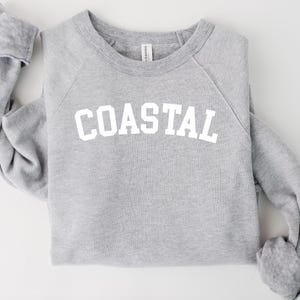 May include: A heather gray sweatshirt with the word "COASTAL" printed in white block letters across the chest. The long-sleeved top is folded on a white surface.