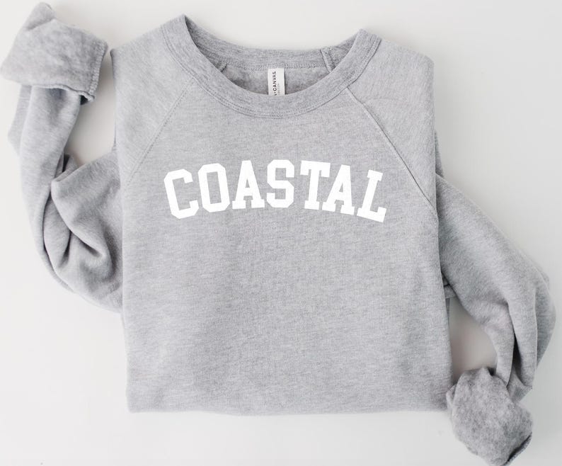 May include: A heather gray sweatshirt with the word "COASTAL" printed in white block letters across the chest. The long-sleeved top is folded on a white surface.
