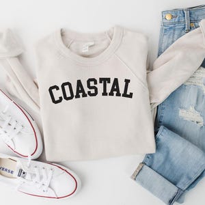 May include: A cream-colored sweatshirt with the word "COASTAL" in black block letters is laid flat next to a pair of light-wash, distressed jeans and white canvas sneakers with red trim. The outfit is arranged on a white surface.