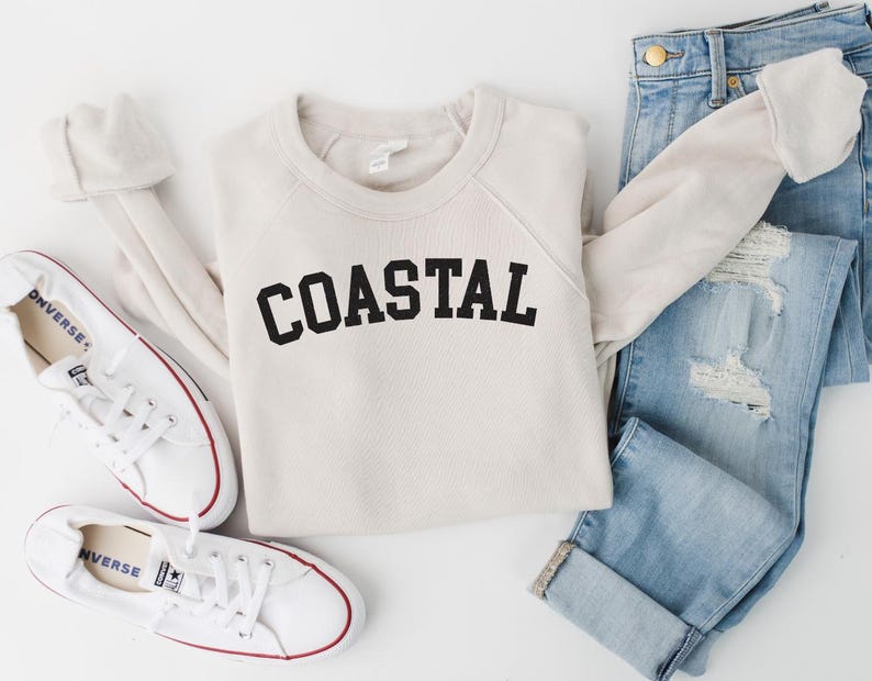 May include: A cream-colored sweatshirt with the word "COASTAL" in black block letters is laid flat next to a pair of light-wash, distressed jeans and white canvas sneakers with red trim. The outfit is arranged on a white surface.