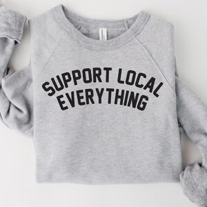 May include: A heather gray sweatshirt with the text "SUPPORT LOCAL EVERYTHING" in bold black letters. The long-sleeved top is folded neatly, showcasing the crew neck and raglan sleeves. The shirt is made of a soft material.
