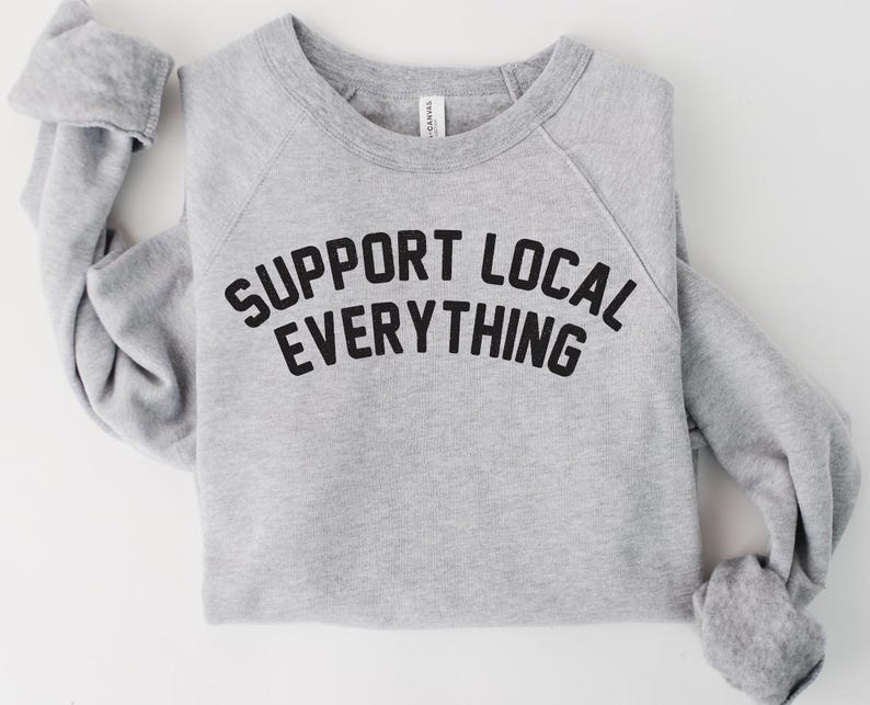 May include: A heather gray sweatshirt with the text "SUPPORT LOCAL EVERYTHING" in bold black letters. The long-sleeved top is folded neatly, showcasing the crew neck and raglan sleeves. The shirt is made of a soft material.