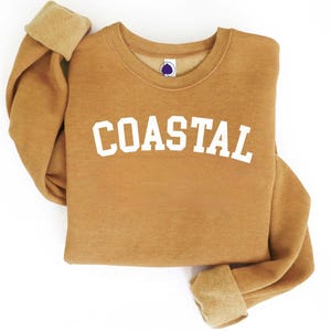 May include: A tan sweatshirt with the word "COASTAL" in white block letters. The sweatshirt has a crew neck and ribbed cuffs. The cuffs are folded over, revealing a lighter tan color.