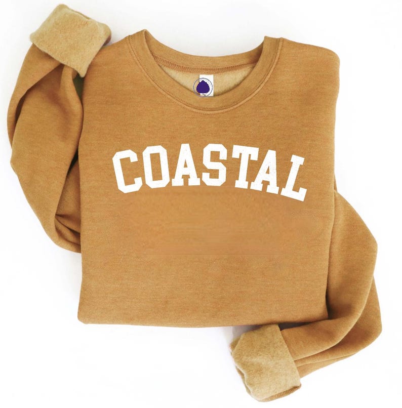 May include: A tan sweatshirt with the word "COASTAL" in white block letters. The sweatshirt has a crew neck and ribbed cuffs. The cuffs are folded over, revealing a lighter tan color.