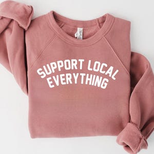 May include: A dusty rose-colored sweatshirt with the text "SUPPORT LOCAL EVERYTHING" in white block letters. The sweatshirt has a crew neck and raglan sleeves. The cuffs are folded up.