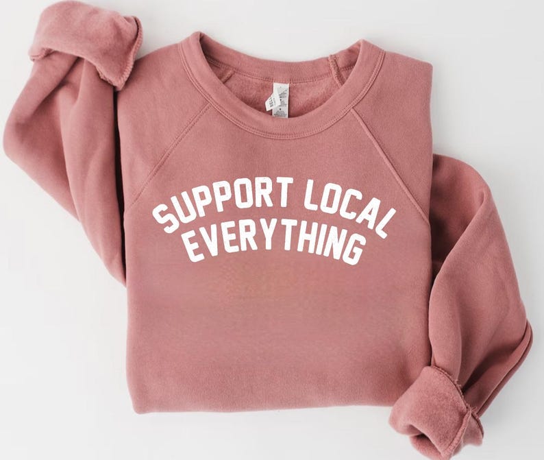 May include: A dusty rose-colored sweatshirt with the text "SUPPORT LOCAL EVERYTHING" in white block letters. The sweatshirt has a crew neck and raglan sleeves. The cuffs are folded up.