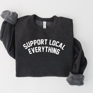 May include: Dark gray sweatshirt with the text "SUPPORT LOCAL EVERYTHING" in white block letters. The sweatshirt has a crew neck and long sleeves with gray cuffs. The garment is laid flat on a white surface.