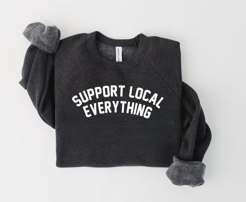 May include: Dark gray sweatshirt with the text "SUPPORT LOCAL EVERYTHING" in white block letters. The sweatshirt has a crew neck and long sleeves with gray cuffs. The garment is laid flat on a white surface.