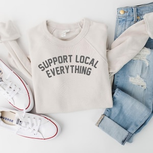 May include: A flat lay featuring a cream-colored sweatshirt with the text "SUPPORT LOCAL EVERYTHING" in gray, paired with light-wash denim jeans and white sneakers. The sneakers have red accents and are laid out with the sweatshirt.