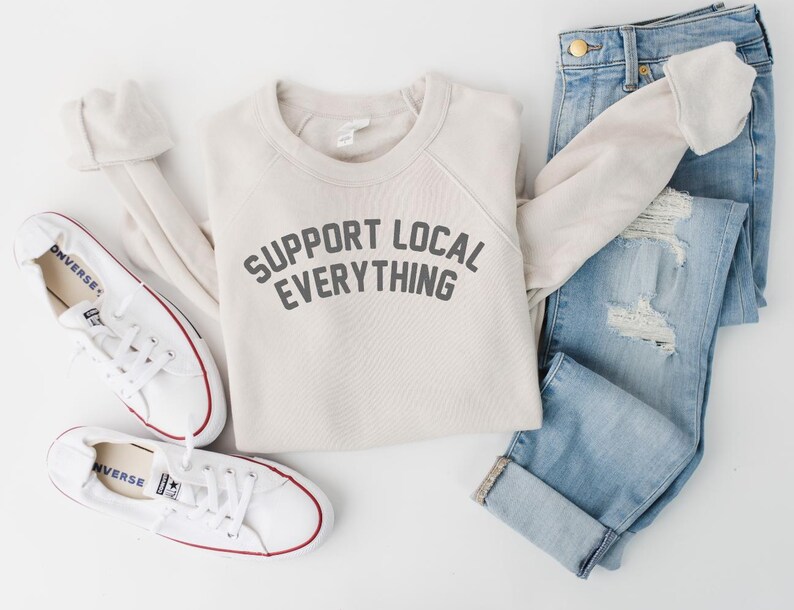 May include: A flat lay featuring a cream-colored sweatshirt with the text "SUPPORT LOCAL EVERYTHING" in gray, paired with light-wash denim jeans and white sneakers. The sneakers have red accents and are laid out with the sweatshirt.