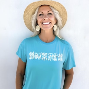 May include: A woman wearing a light blue t-shirt with a white folk art design featuring a rooster, flowers, and people. She is wearing a straw hat and large oval earrings.