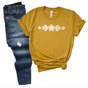 May include: A mustard yellow t-shirt with a white floral design across the chest. The shirt is shown with a pair of blue jeans.