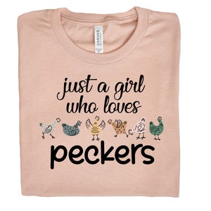 May include: A light pink t-shirt with the text "just a girl who loves peckers" in black. There are six cartoon chickens in a row below the text.