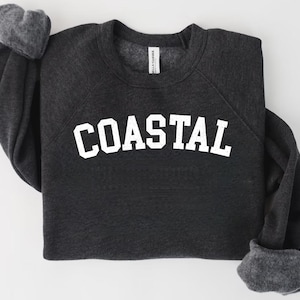 May include: Dark gray sweatshirt with the word "COASTAL" in large, white, block letters across the chest. The sweatshirt has a crew neck and long sleeves with gray cuffs. The fabric appears soft and comfortable.