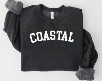 May include: Dark gray sweatshirt with the word "COASTAL" in large, white, block letters across the chest. The sweatshirt has a crew neck and long sleeves with gray cuffs. The fabric appears soft and comfortable.