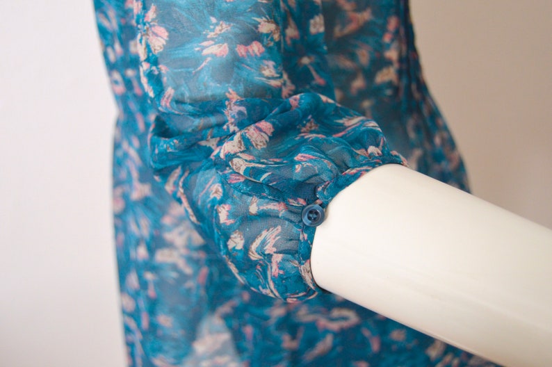 K&ouml;nnte beinhalten: Eine Bluse mit blauem und rosafarbenem Blumendruck und einem Knopfdetail am &Auml;rmel.