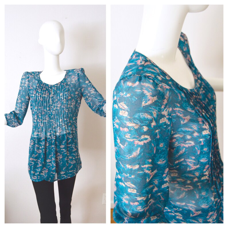 K&ouml;nnte beinhalten: Eine durchsichtige Bluse in Tealblau mit Blumenmuster. Die Bluse hat einen runden Ausschnitt und lange &Auml;rmel. Der Stoff ist halbtransparent und hat ein zartes, flie&szlig;endes Gef&uuml;hl.