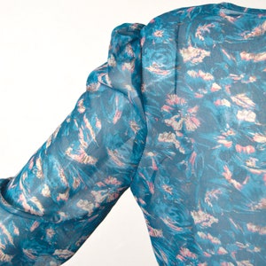 K&ouml;nnte beinhalten: Eine blaue, durchsichtige Bluse mit einem rosa-wei&szlig;en Blumendruck. Die Bluse hat einen Puff&auml;rmel und eine lockere Passform.