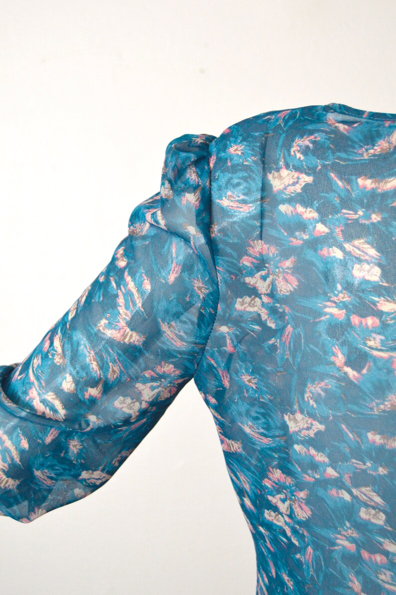 K&ouml;nnte beinhalten: Eine blaue, durchsichtige Bluse mit einem rosa-wei&szlig;en Blumendruck. Die Bluse hat einen Puff&auml;rmel und eine lockere Passform.