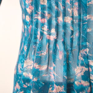 K&ouml;nnte beinhalten: Eine durchsichtige Bluse in Tealblau mit einem Blumendruck in Rosa- und Wei&szlig;t&ouml;nen. Die Bluse hat vertikale Falten auf der Vorderseite.