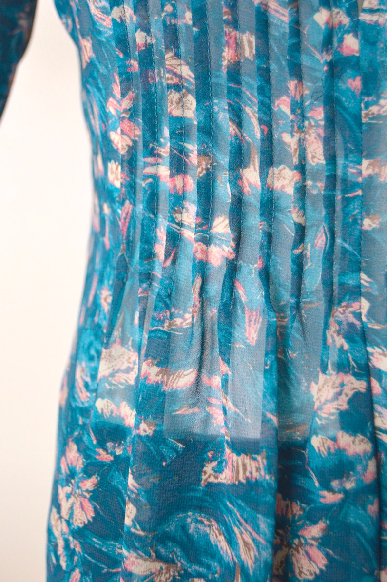 K&ouml;nnte beinhalten: Eine durchsichtige Bluse in Tealblau mit einem Blumendruck in Rosa- und Wei&szlig;t&ouml;nen. Die Bluse hat vertikale Falten auf der Vorderseite.