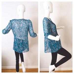 K&ouml;nnte beinhalten: Eine durchsichtige blaue Bluse mit einem rosafarbenen Blumenmuster. Die Bluse hat einen V-Ausschnitt und 3/4 &Auml;rmel.
