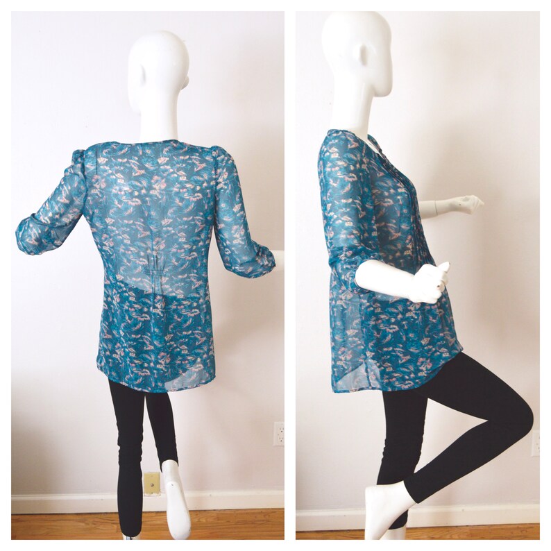 K&ouml;nnte beinhalten: Eine durchsichtige blaue Bluse mit einem rosafarbenen Blumenmuster. Die Bluse hat einen V-Ausschnitt und 3/4 &Auml;rmel.