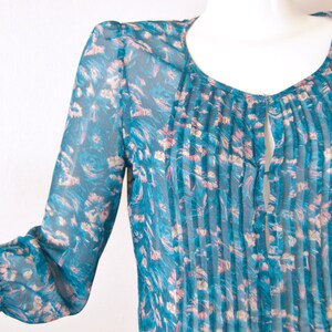 K&ouml;nnte beinhalten: Eine durchsichtige Bluse in Tealblau mit Blumendruck und einer gefalteten Vorderseite. Die Bluse hat einen Schl&uuml;ssellochausschnitt am Halsausschnitt.