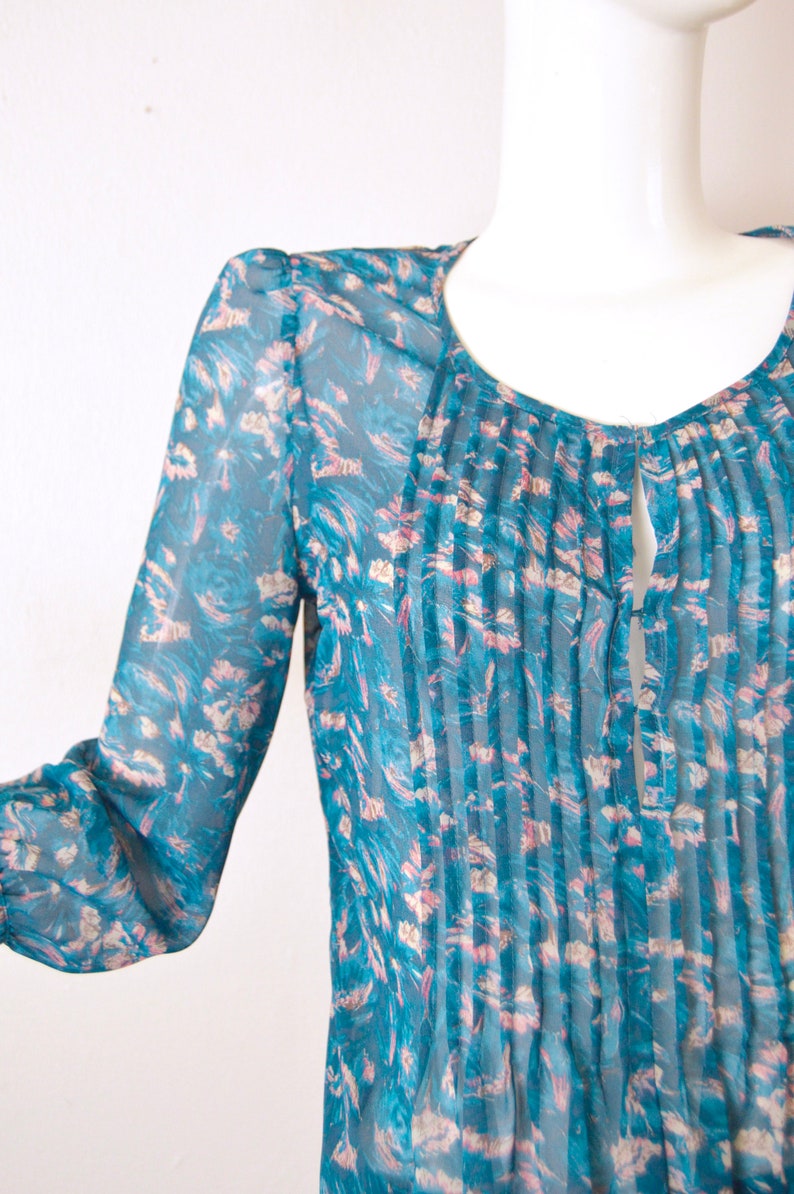 K&ouml;nnte beinhalten: Eine durchsichtige Bluse in Tealblau mit Blumendruck und einer gefalteten Vorderseite. Die Bluse hat einen Schl&uuml;ssellochausschnitt am Halsausschnitt.