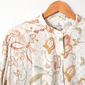 May include: A white button-up shirt with a floral print in shades of brown, pink, and green. The shirt has a mandarin collar and a single button closure. The label reads "Liz Claiborne Collection".