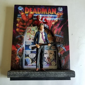 Peut inclure: Une vitrine de bande dessinée DC Comics Deadman avec une figurine d'un homme tenant une arme à feu. La figurine est debout devant une couverture de bande dessinée avec le titre "Deadman" et un prix de 3,99 $. La vitrine est en bois et a une base noire.