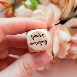 Op de afbeelding: Een kleine houten kubus met de woorden "you're amazing" erop gegraveerd. De kubus wordt in een hand gehouden.