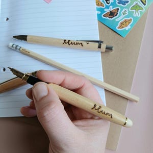 Puede incluir: Dos bolígrafos de madera con la palabra "Mum" grabada. Uno es un bolígrafo y el otro es una pluma estilográfica.
