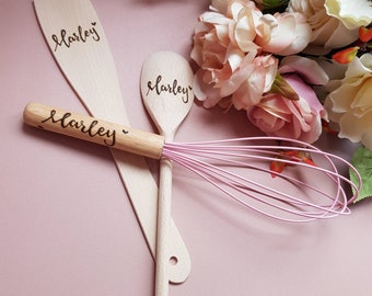 May include: A set of personalized wooden kitchen utensils, including a spatula, spoon, and whisk. Each utensil is engraved with the name "Marley" in a cursive font. The whisk features a pink wire head. The background includes pink flowers.