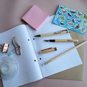 Puede incluir: Un bloc de notas rosa, un cuaderno con páginas rayadas, un rollo de cinta adhesiva, un lápiz de madera, un bolígrafo de madera, una pluma estilográfica y una hoja de pegatinas con diseños de mariposas. El bolígrafo y el lápiz están grabados con la palabra "Mamá".