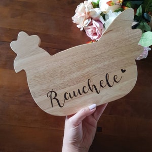 May include: A wooden cutting board shaped like a chicken, held by a person. The board has a light wood grain and features the name "Rauchele" in a cursive font, with a small heart symbol. Decorative flowers are in the background.