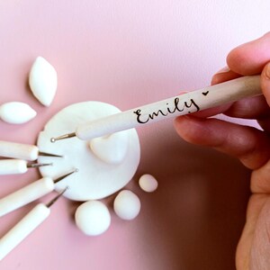 May include: A close-up of a wooden tool with the name "Emily" engraved on it. The tool is being used to shape white clay. There are several other tools and pieces of clay in the background.