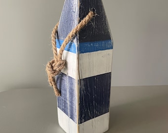 May include: A decorative wooden buoy painted with navy blue and white stripes, accented with a light blue stripe. A natural rope is tied around the top. The buoy is a nautical-themed decorative item.