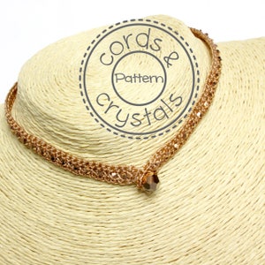 May include: A gold-toned necklace with a crystal pendant. The necklace is made of braided cord and features a delicate, intricate design. The pendant is a faceted crystal with a rose gold tone.