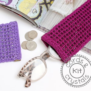 Peut inclure: Deux étuis à lunettes en crochet, l'un violet et l'autre rose, avec des embellissements en cristal. Les étuis sont posés sur une surface blanche avec une paire de lunettes et des pièces de monnaie.