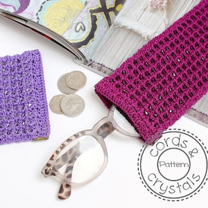 Könnte beinhalten: Zwei gehäkelte Brillenetuis, eines lila und eines rosa, mit schwarzen Perlen. Das rosa Etui liegt auf einer weißen Oberfläche mit einer Brille und einem Magazin. Das Magazin hat den Text "cords & crystals pattern" auf dem Cover.