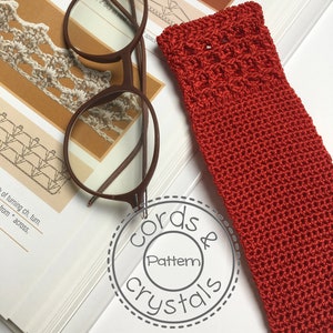 Könnte beinhalten: Nahaufnahme eines roten Häkel-Lesezeichens mit dem Text "Cords & Crystals Pattern" in einem Kreis. Das Lesezeichen liegt auf einem Buch mit einer Brille darauf.