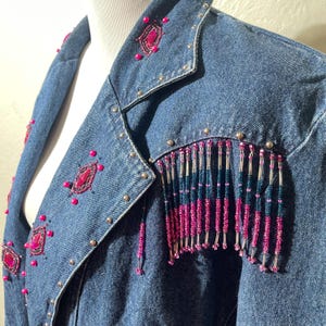 Pode incluir: Uma jaqueta jeans azul adornada com detalhes rosa e prateados. A jaqueta apresenta franjas de contas rosa e miçangas decorativas rosa e prateadas. Tachas prateadas são colocadas ao longo da gola e dos ombros. A jaqueta é exibida em um manequim.