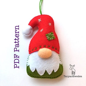 May include: A felt Christmas gnome ornament with a red hat, white beard, and green body. The gnome has a small white pom-pom on the top of its hat and a green gift box on the front. The ornament is hanging from a green string.