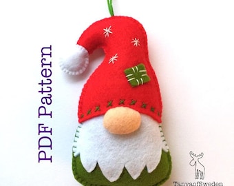 May include: A felt Christmas gnome ornament with a red hat, white beard, and green body. The gnome has a small white pom-pom on the top of its hat and a green gift box on the front. The ornament is hanging from a green string.