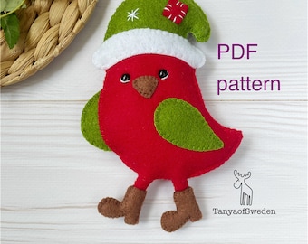 Puede incluir: Adorno de pájaro de fieltro hecho a mano en rojo, verde y marrón, con un gorro verde con borde blanco. El pájaro tiene botas marrones y una cuerda roja para colgar. El texto "PDF pattern" es visible.